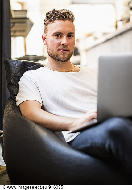 Portrait of confident young businessman using laptop while sitting on bean bag chair in new office