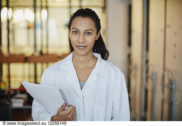 Portrait of confident young chemistry student wearing lab coat standing with book in university
