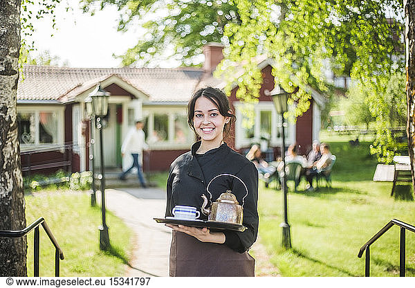 Portrait of confident young female waitress smiling while holding serving tray against outdoor cafe