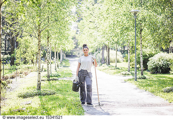 Portrait of confident young woman holding watering can and rake on footpath at garden