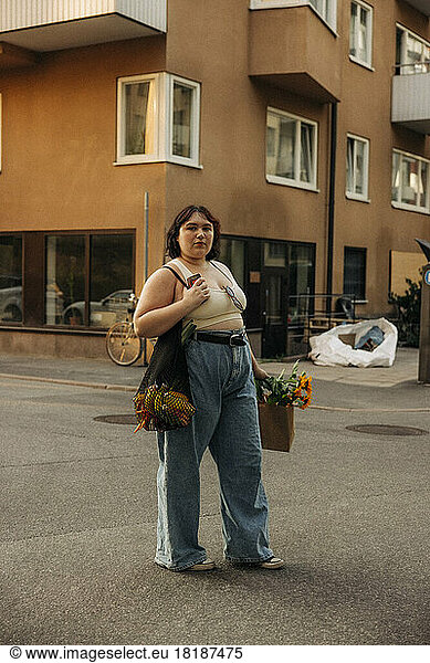 Portrait of confident young woman with shopping bag standing against building