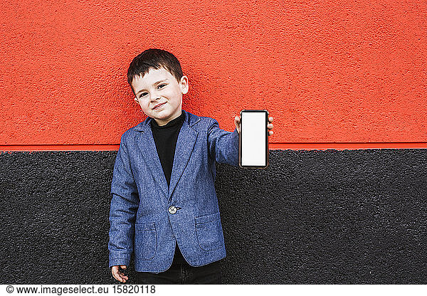 Portrait of content little boy wearing suit coat showing smartphone