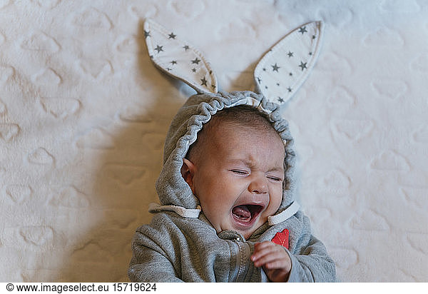 Portrait of crying baby girl in a rabbit hoodie lying on white blanket