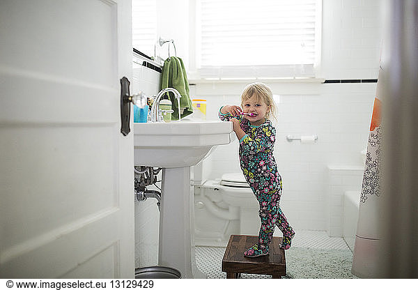 Portrait of cute baby girl brushing teeth while standing on stool in bathroom