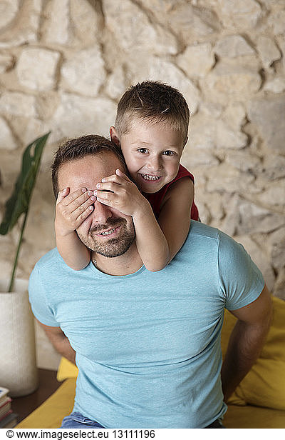 Portrait of cute smiling son covering father's eyes with hands while playing at home