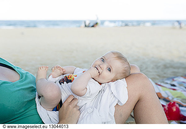 Portrait of cute son with fingers in mouth lying on mother's laps at beach