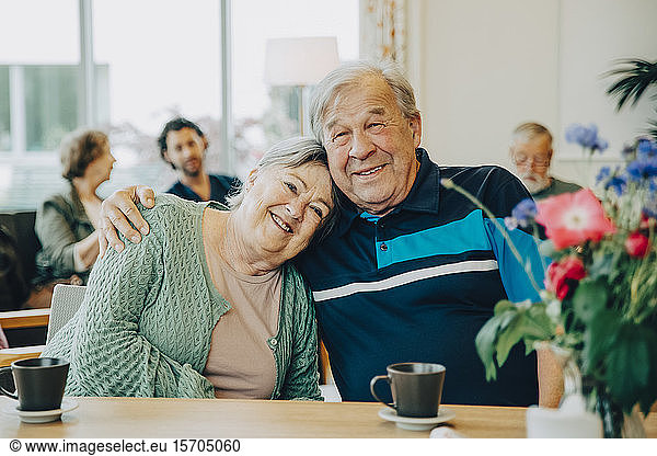 Portrait of elderly senior friends sitting with arm around at dining table in nursing home