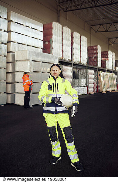 Portrait of female blue-collar worker in protective workwear holding hardhat while standing at industry