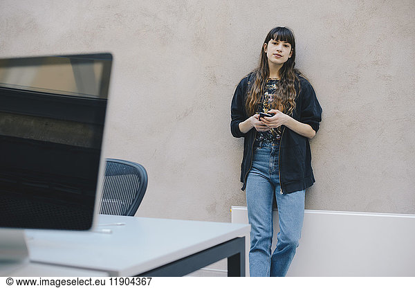 Portrait of female computer programmer holding smart phone while standing against beige wall in office