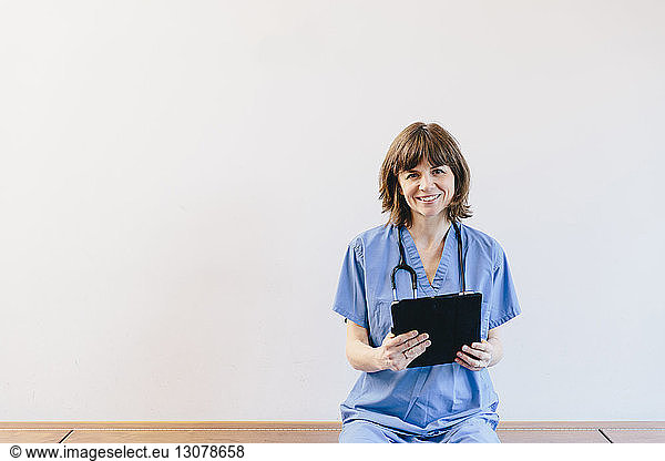 Portrait of female doctor holding tablet computer while sitting on bench against white background