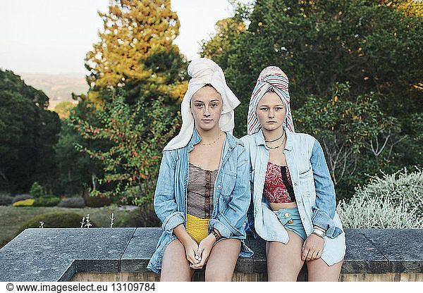 Portrait of female friends with hair wrapped in towel sitting on retaining wall