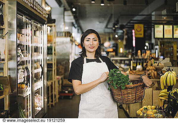 Portrait of female retail clerk carrying wicker basket while standing at organic grocery store