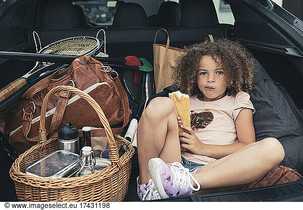 Portrait of girl eating bread while sitting with luggage in car trunk