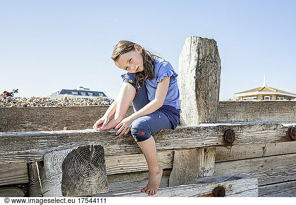 Portrait of girl (4-5) sitting on wooden structure on beach
