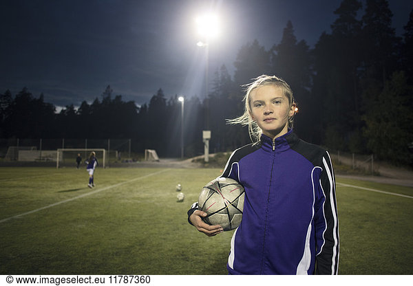 Portrait of girl standing with soccer ball on field against trees at night