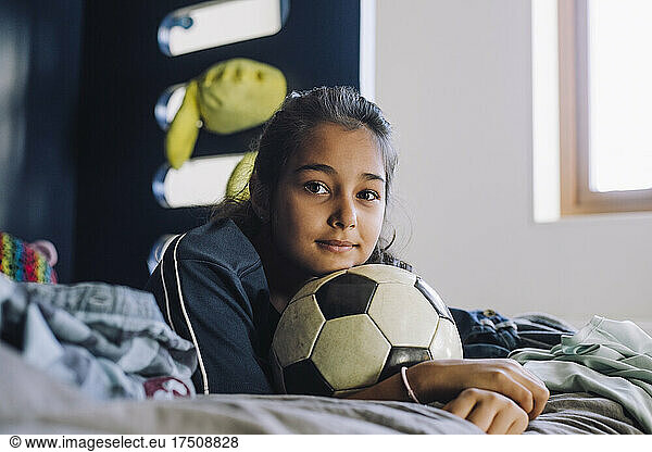 Portrait of girl with soccer ball lying on bed at home