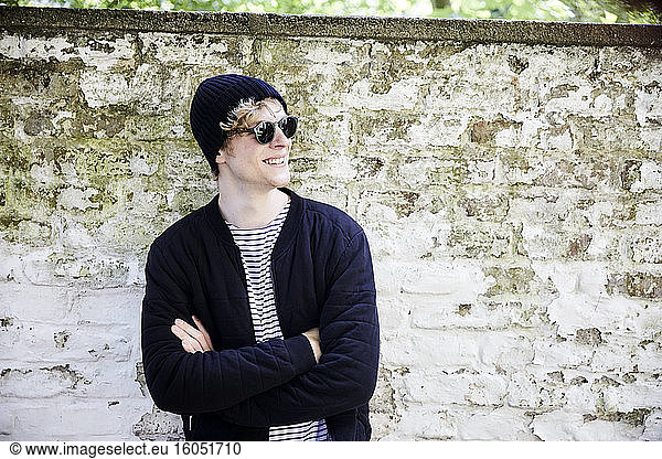 Portrait of grinning young man with headphones wearing sunglasses and cap in front of weathered wall