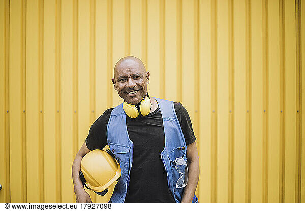 Portrait of happy bald construction worker with hardhat and ear protectors in front of yellow metal wall