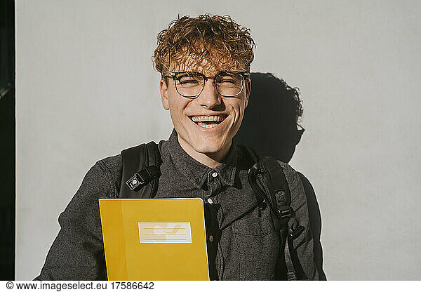 Portrait of happy blond male student wearing eyeglasses holding book against gray wall