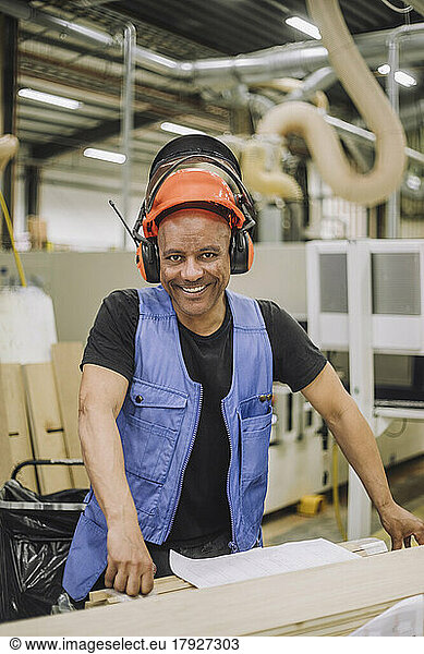 Portrait of happy carpenter wearing hardhat and ear protectors working in warehouse