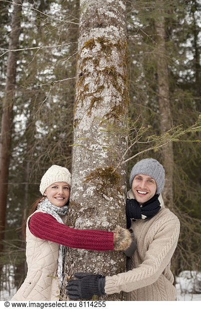 Portrait of happy couple hugging tree trunk in woods