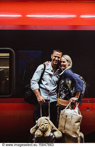 Portrait of happy couple with luggage standing against train at railroad station