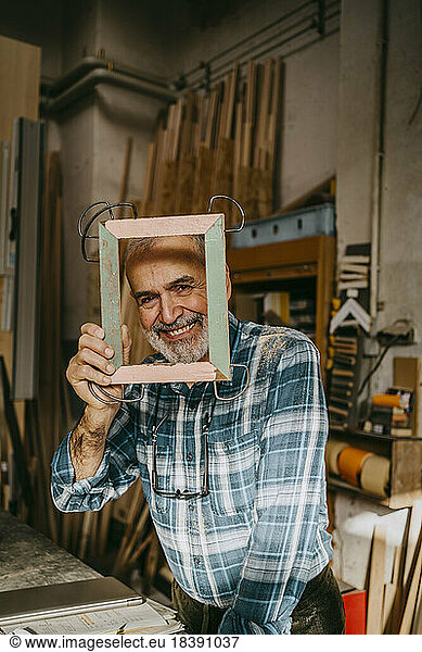 Portrait of happy craftsman looking through picture frame at workshop