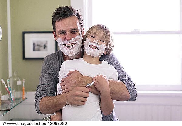 Portrait of happy father and son with shaving cream on face standing in bathroom