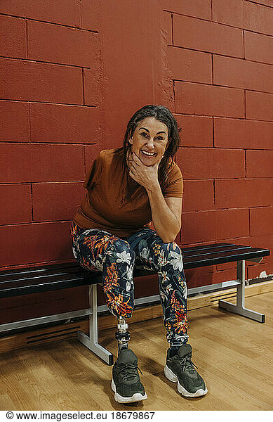 Portrait of happy female athlete with amputated leg sitting on bench at sports court