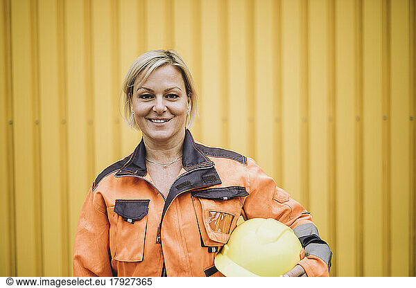 Portrait of happy female construction worker with hardhat against yellow metal wall