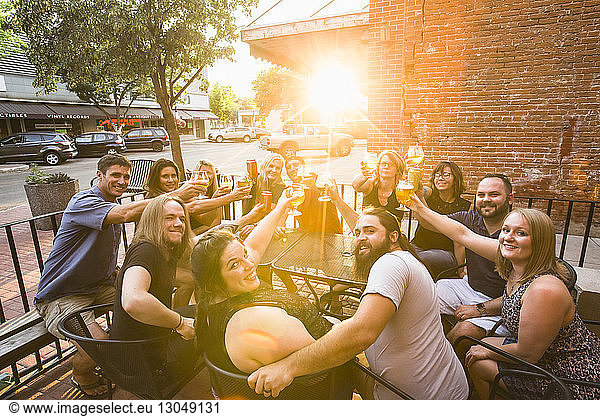 Portrait of happy friends toasting beer while sitting at sidewalk cafe during sunny day