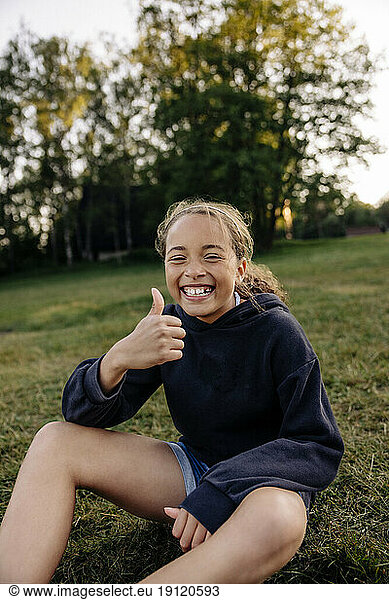 Portrait of happy girl showing thumbs up while sitting on grass in playground