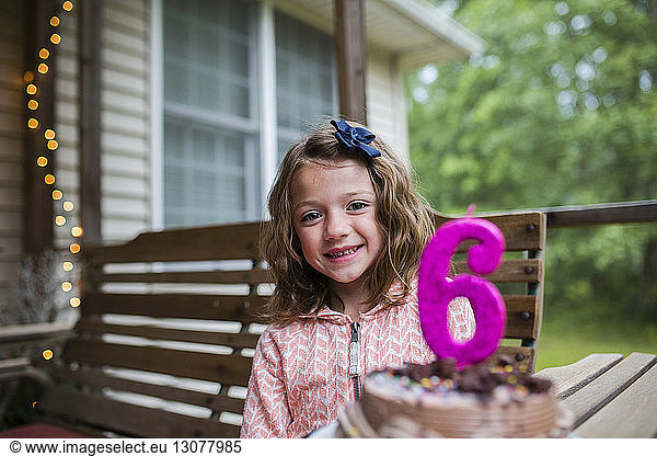 Portrait of happy girl sitting by birthday cake with number 6 candle at porch