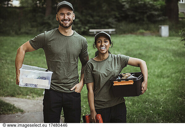 Portrait of happy male and female technician coworkers with toolboxes at back yard