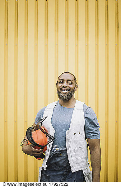 Portrait of happy mature construction worker standing with hardhat against yellow metal wall