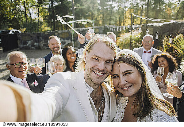 Portrait of happy newlywed couple taking selfie with guests in background on wedding day