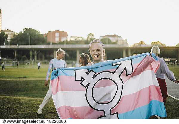 Portrait of happy non-binary person standing with transgender symbol on flag at park