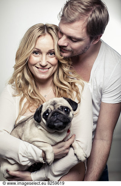Portrait of happy pregnant woman holding pug dog with man embracing her from behind