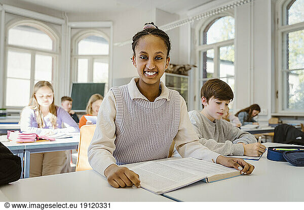 Portrait of happy schoolgirl sitting at desk with male friend in classroom