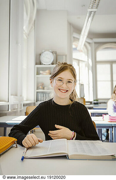 Portrait of happy schoolgirl with book at desk in classroom