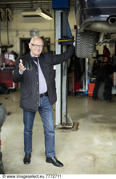 Portrait of happy senior man gesturing while standing by car jack in auto repair shop
