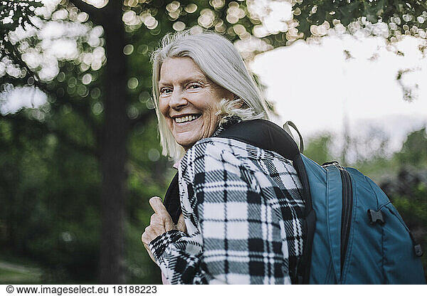 Portrait of happy senior woman looking over shoulder carrying backpack while hiking