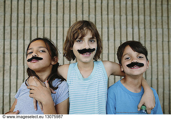 Portrait of happy siblings with artificial mustache against wall