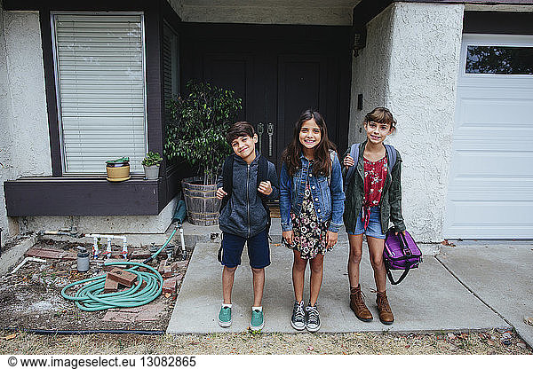 Portrait of happy siblings with backpack standing in front of house