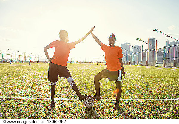 Portrait of happy sportswomen giving high-five with soccer ball on field during sunny day