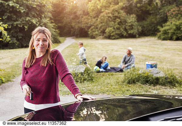 Portrait of happy woman standing by electric car with family in background at park