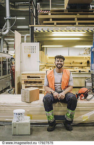 Portrait of happy young carpenter wearing reflective clothing sitting by hardhat in warehouse