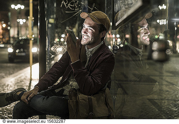 Portrait of happy young man on the phone sitting at bus stop by night  Lisbon  Portugal