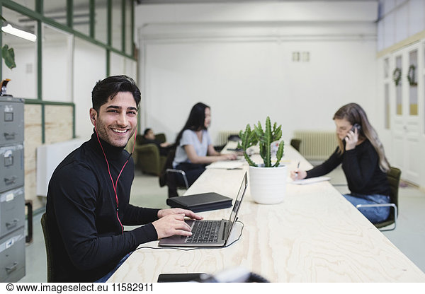 Portrait of happy young man sitting at desk with colleagues working in background