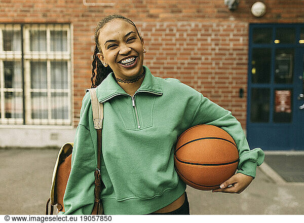 Portrait of happy young woman holding basketball under arm while standing in front of building